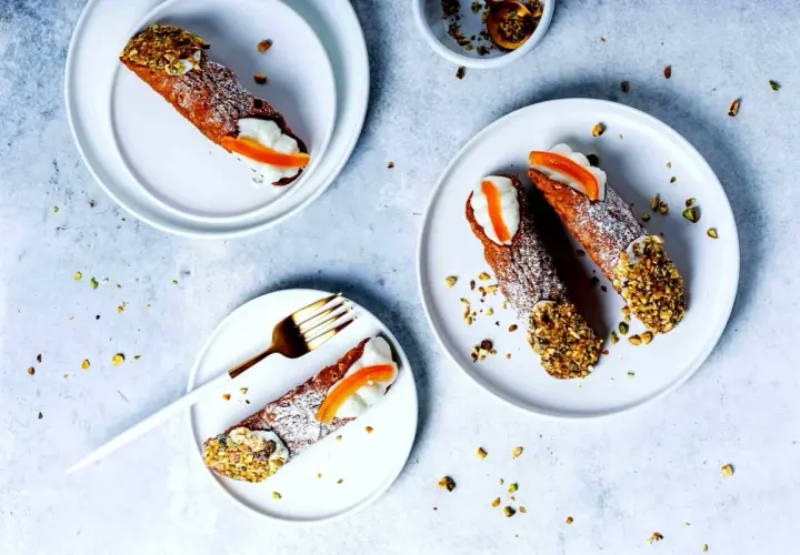 Tre cannoli siciliani, guarniti con crema, arancia candita e pistacchi tritati, su piatti bianchi, spolverati di zucchero a velo.