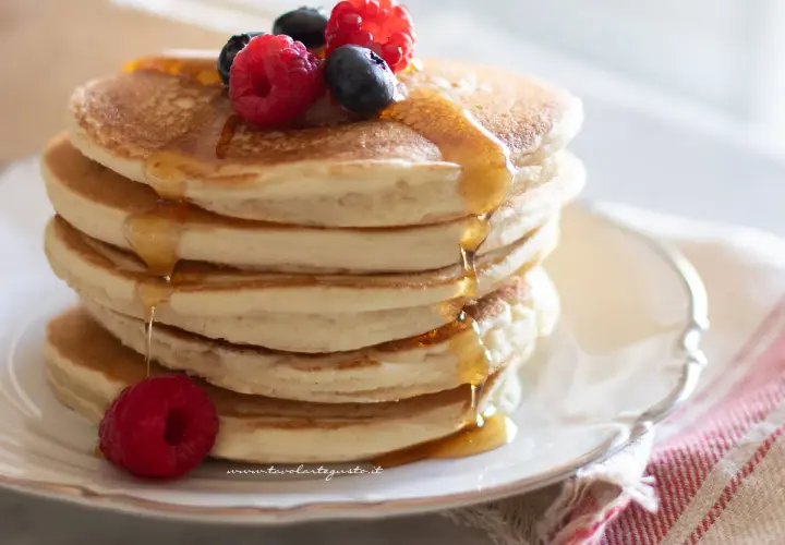 Pila di soffici pancake senza glutine, guarniti con frutti di bosco e sciroppo d'acero che cola.
