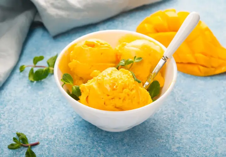 Coppa di sorbetto al mango con menta fresca e una fetta di mango sullo sfondo.