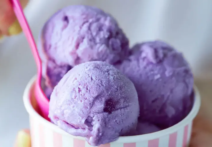 Tre palline di gelato viola in una coppetta a righe rosa e bianche, con un cucchiaino rosa. Un dolce piacere che non causa stitichezza.