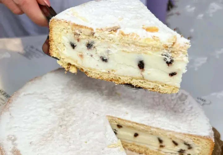 Una fetta di cassata al forno, ricca di crema e gocce di cioccolato, viene sollevata da una torta intera cosparsa di zucchero a velo.