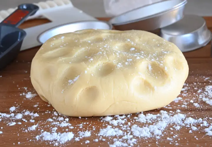 Impasto di pasta frolla pronto per essere lavorato, cosparso di zucchero a velo su un tagliere di legno. Attrezzi da cucina sullo sfondo.