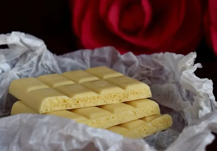 Barrette di cioccolato bianco, con la loro texture ariosa, avvolte in carta crespa, sullo sfondo di una rosa rossa. Ecco com'è fatto il cioccolato bianco.