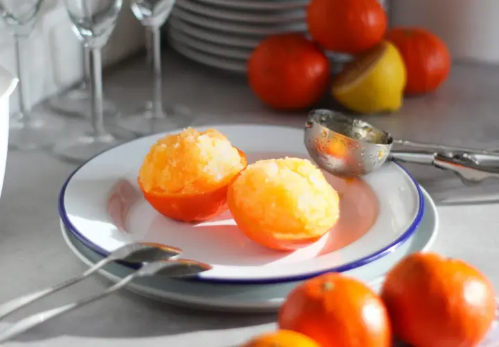 Sorbetto al mandarino senza gelatiera servito in coppette di mandarino. Frutta fresca e cucchiaio da gelato sullo sfondo.