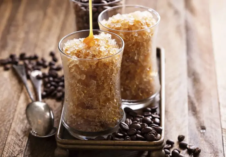 Due bicchieri di granita al caffè, con sciroppo che cola su uno. Chicchi di caffè tostati e cucchiaini completano la scena.