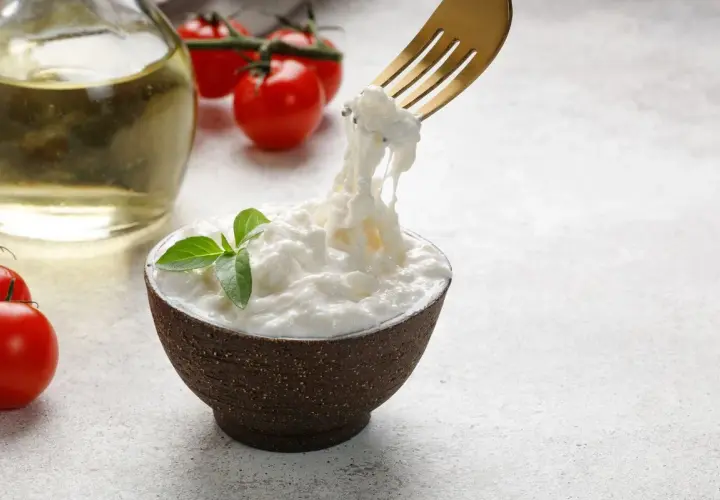 Una ciotola di stracciatella fatta in casa, con pomodorini e olio d'oliva sullo sfondo. Una forchetta solleva fili di formaggio fresco.