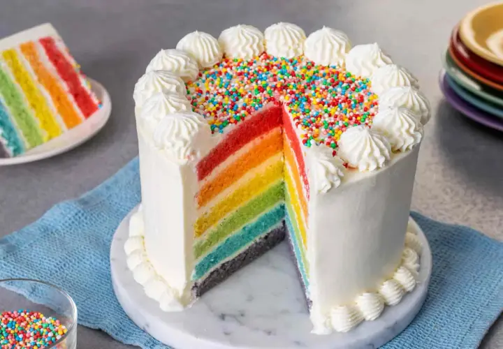 Torta arcobaleno con strati colorati, glassa bianca e zuccherini. Una fetta mostra i colori vivaci, come un vero arcobaleno.