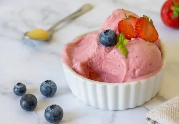 Delizioso gelato rosa, guarnito con mirtilli e fragole, servito in una ciotola bianca. Intorno, altri mirtilli e un cucchiaino con un po' di miele. Perfetto per un momento di fresco piacere, questo gelato palline di ghiaccio è un sogno.
