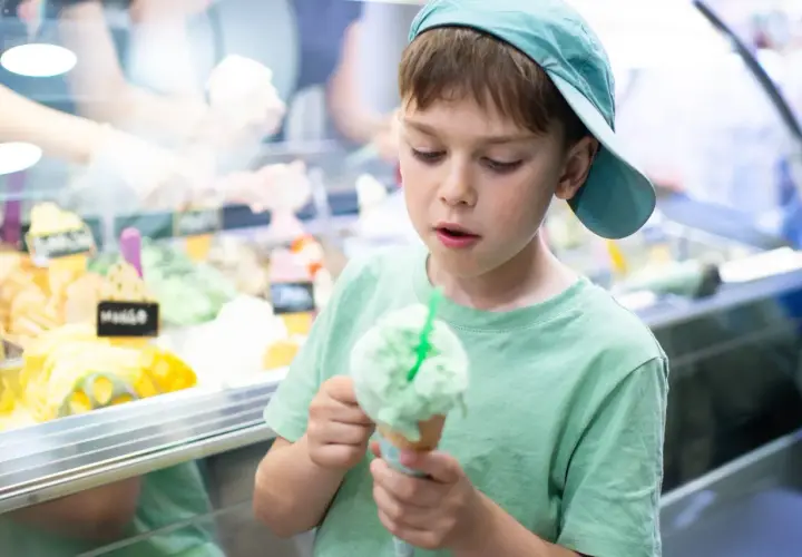 Bambino con cappellino azzurro gusta un gelato verde, pensando a chi ha inventato questa delizia.
