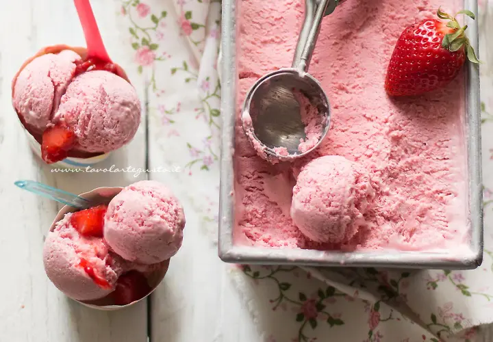 Coppette e vaschetta piene di cremoso gelato alla fragola, con fragole fresche e un cucchiaio pronto per servire.
