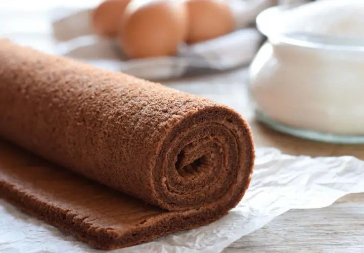 Rotolo di pasta biscotto al cacao, pronto per essere farcito. Ingredienti freschi come uova e zucchero sullo sfondo.