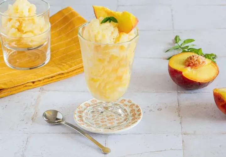 Rinfrescante granita alla pesca, guarnita con una fetta di pesca e menta. Accanto, pesche fresche e un cucchiaino.