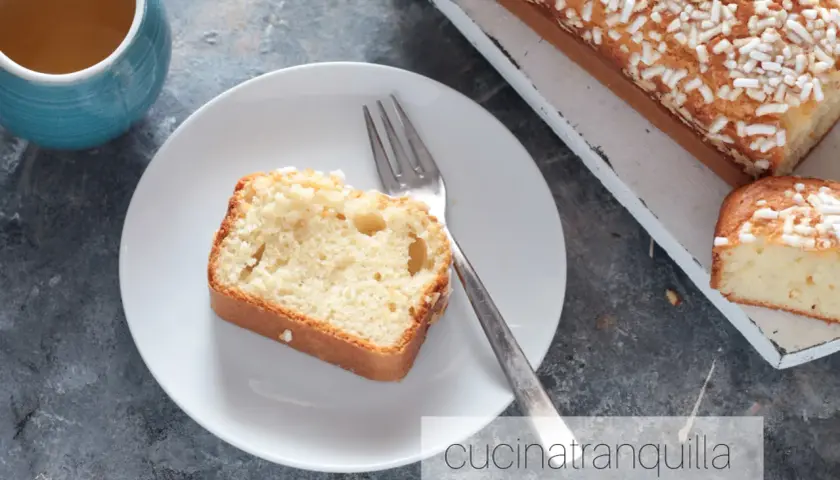 Una fetta di plumcake yogurt greco, soffice e cosparso di zuccherini, attende su un piatto bianco, accanto a una tazza di tè.