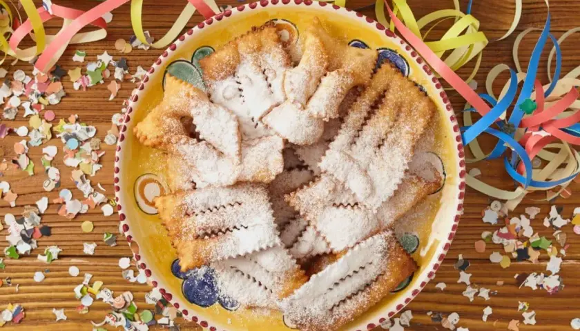 Chiacchiere di Carnevale Benedetta Rossi, cosparse di zucchero a velo, su un piatto decorato, circondate da coriandoli e stelle filanti.