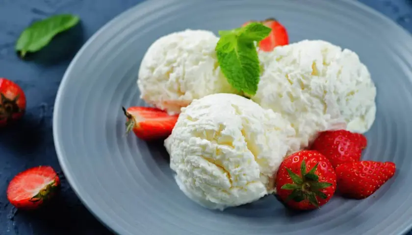Tre palline di cremoso gelato alla ricotta, guarnite con fragole fresche e una fogliolina di menta, su un piatto grigio.