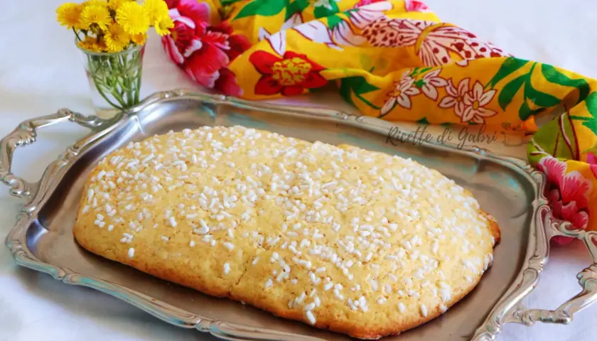 Una ciambella romagnola, cosparsa di zuccherini, su un vassoio d'argento, con fiori gialli e un foulard colorato sullo sfondo.