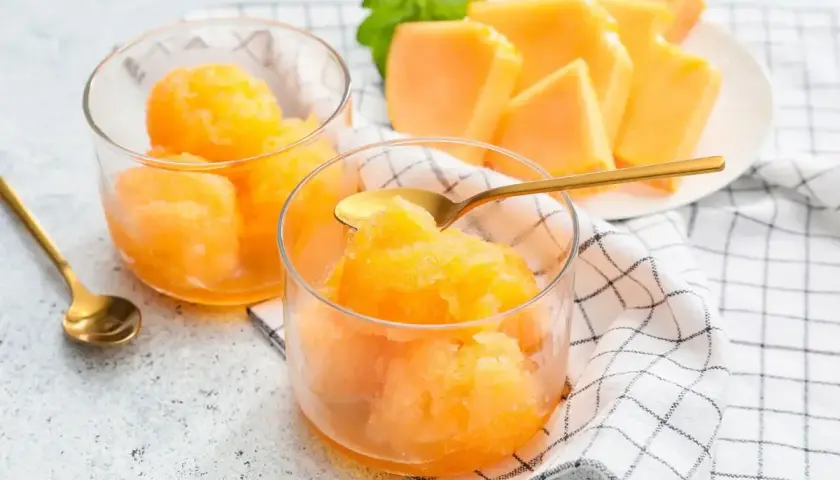 Sorbetto melone fresco e dissetante, servito in coppette di vetro con cucchiaini dorati. Fette di melone e menta sullo sfondo.
