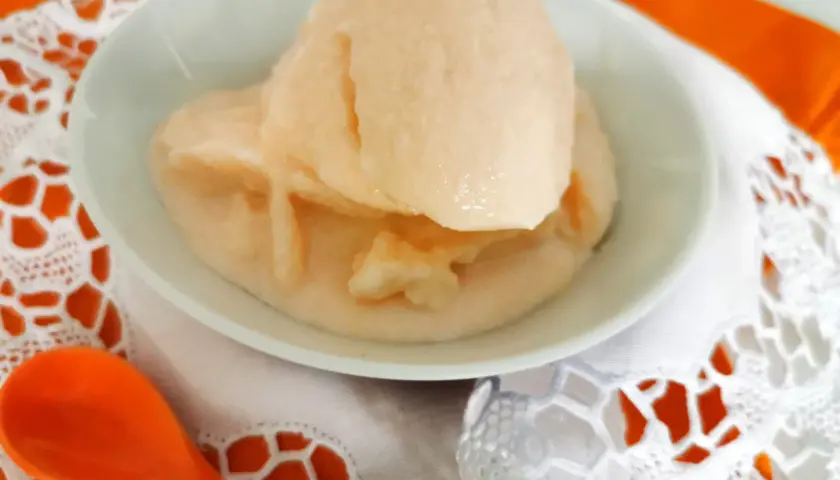 Delizioso gelato al melone solo frutta, servito in una ciotola bianca su un tovagliolo di pizzo bianco e un runner arancione. Un cucchiaio arancione è posizionato a sinistra.