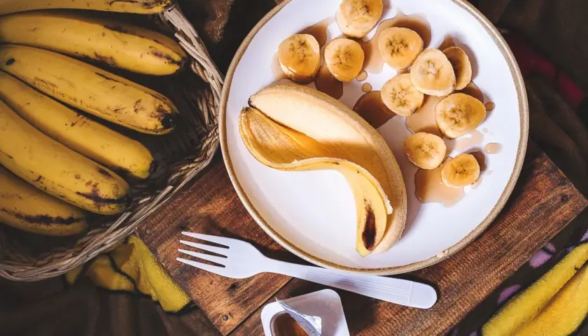Un cestino di banane mature accanto a una banana sbucciata e affettata su un piatto, con sciroppo. Un'idea per sostituire lo zucchero con la banana.