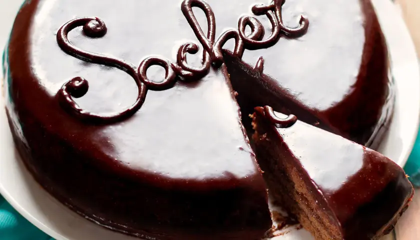 Una lucida torta Sacher, con una fetta tagliata, mostra la sua ricca copertura al cioccolato e la scritta "Sacher" decorata.