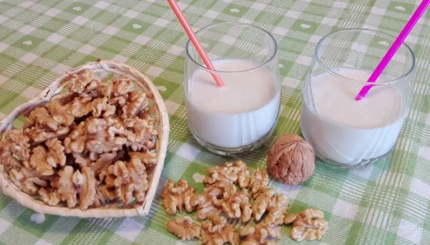 Due bicchieri con latte di noci fatto in casa, una ciotola a forma di cuore piena di noci e noci sparse sul tavolo.
