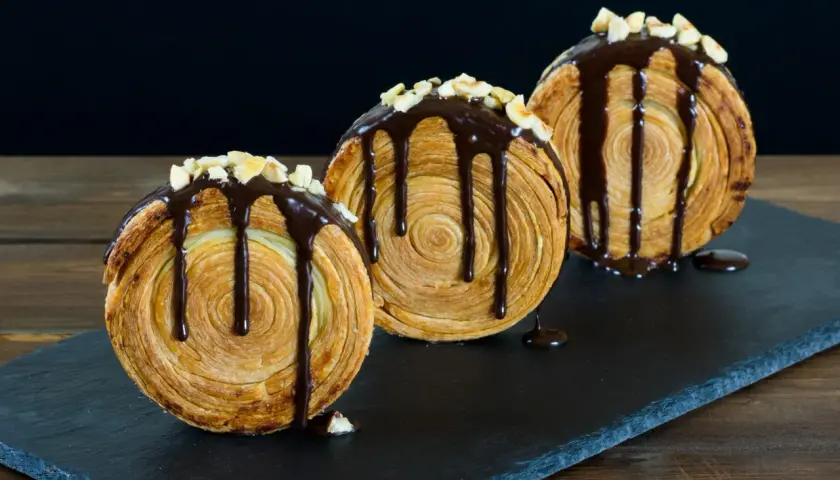 Tre deliziosi new york roll, ricoperti di cioccolato fuso e granella di nocciole, su una lavagna scura.