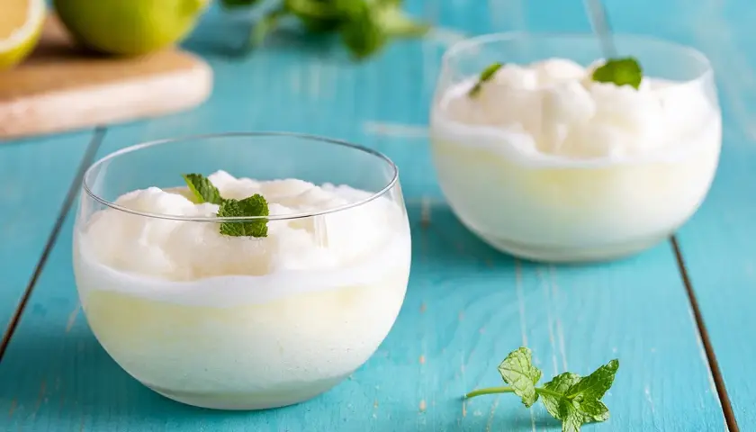 Due bicchieri di sorbetto con albume, guarniti con menta fresca, su un tavolo di legno blu.