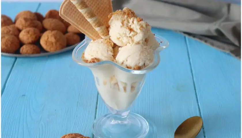 Coppa di gelato amaretto con biscotti e cialde su tavolo blu.