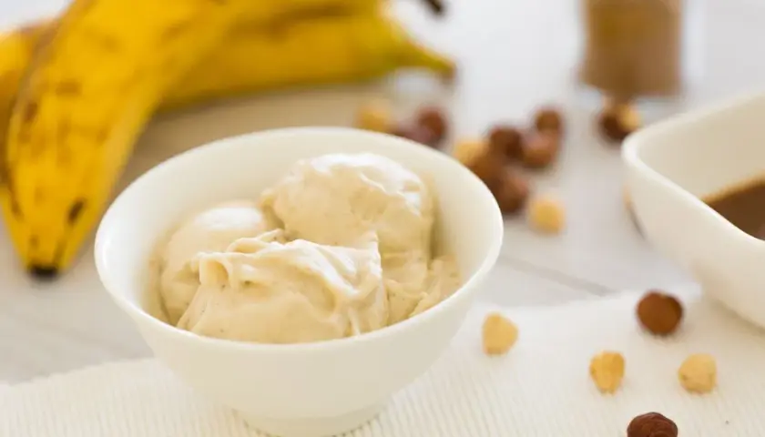 Una ciotola di cremoso gelato alla banana, con banane mature sullo sfondo e nocciole sparse.