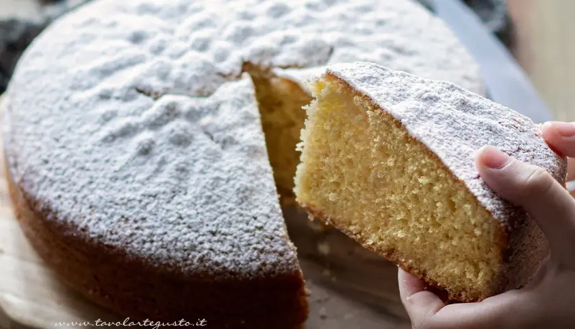 Una fetta di soffice torta margherita, cosparsa di zucchero a velo, viene sollevata da una mano.