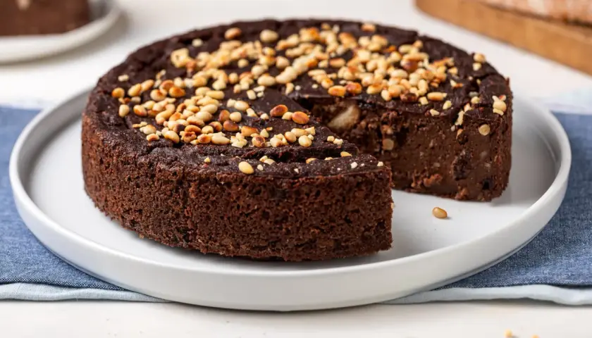 Torta paesana al cioccolato, ricca e umida, guarnita con pinoli tostati. Una fetta è stata già servita, invitando a gustare questo dolce rustico.