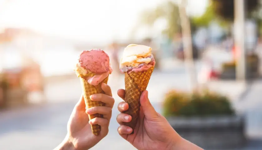 Due mani sollevano due coni gelato, uno rosa e uno con sfumature crema e rosa, celebrando la giornata del gelato.