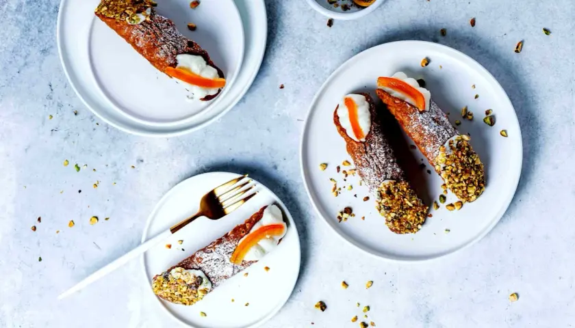 Tre cannoli siciliani, guarniti con crema, arancia candita e pistacchi tritati, su piatti bianchi, spolverati di zucchero a velo.