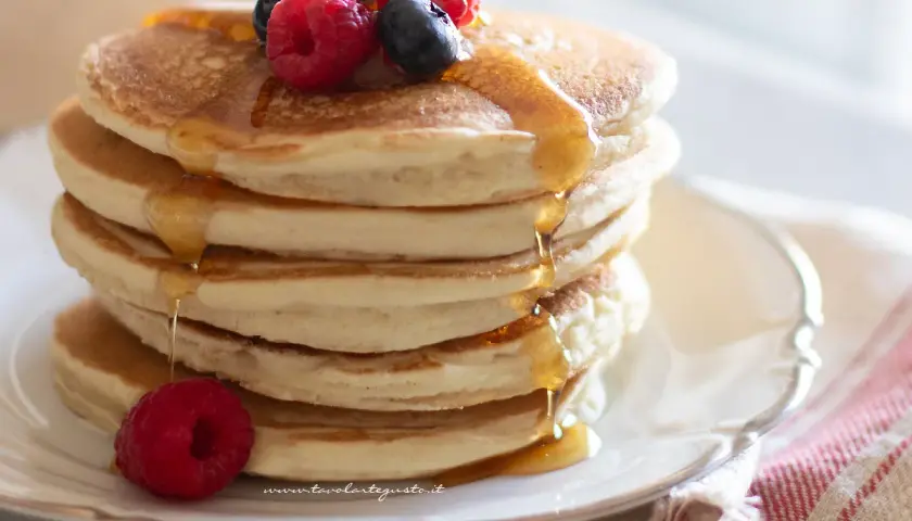 Pila di soffici pancake senza glutine, guarniti con frutti di bosco e sciroppo d'acero che cola.