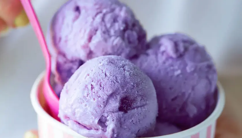 Tre palline di gelato viola in una coppetta a righe rosa e bianche, con un cucchiaino rosa. Un dolce piacere che non causa stitichezza.