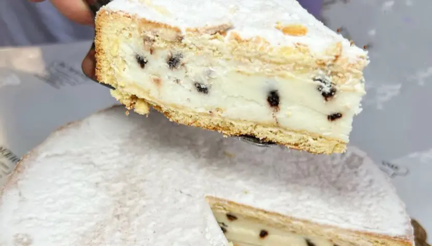 Una fetta di cassata al forno, ricca di crema e gocce di cioccolato, viene sollevata da una torta intera cosparsa di zucchero a velo.