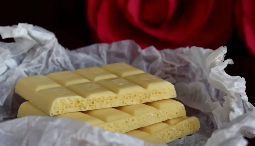 Barrette di cioccolato bianco, con la loro texture ariosa, avvolte in carta crespa, sullo sfondo di una rosa rossa. Ecco com'è fatto il cioccolato bianco.