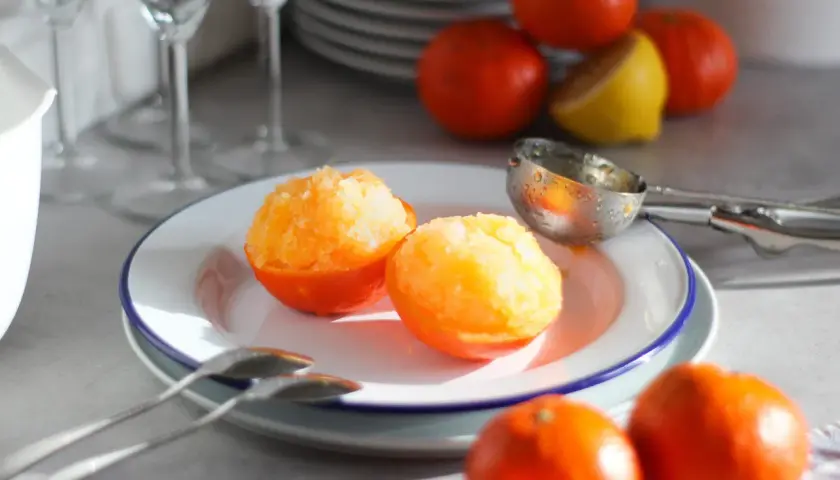 Sorbetto al mandarino senza gelatiera servito in coppette di mandarino. Frutta fresca e cucchiaio da gelato sullo sfondo.