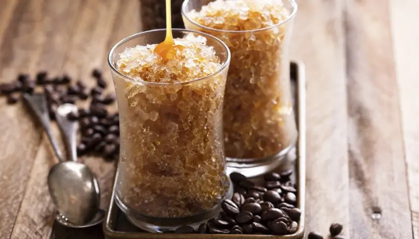 Due bicchieri di granita al caffè, con sciroppo che cola su uno. Chicchi di caffè tostati e cucchiaini completano la scena.