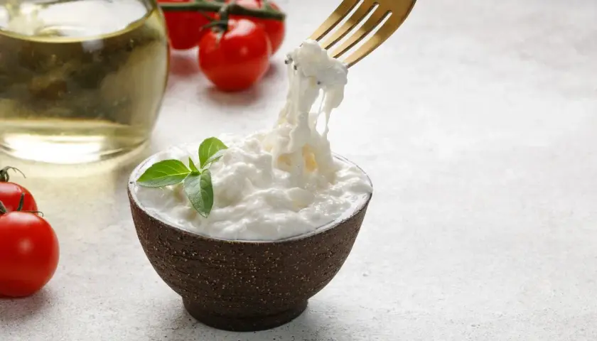 Una ciotola di stracciatella fatta in casa, con pomodorini e olio d'oliva sullo sfondo. Una forchetta solleva fili di formaggio fresco.