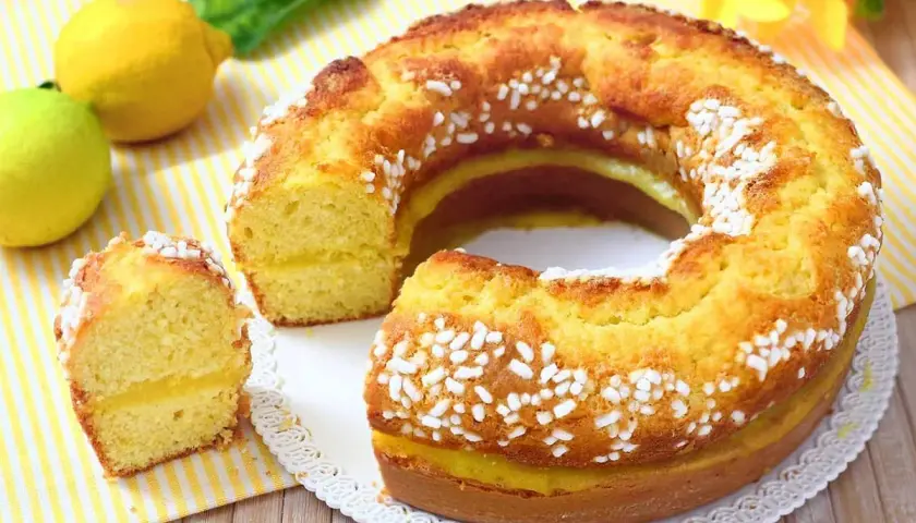 Ciambellone al limone soffice e profumato, con glassa gialla e zuccherini bianchi. Due limoni freschi e una foglia verde completano la scena.
