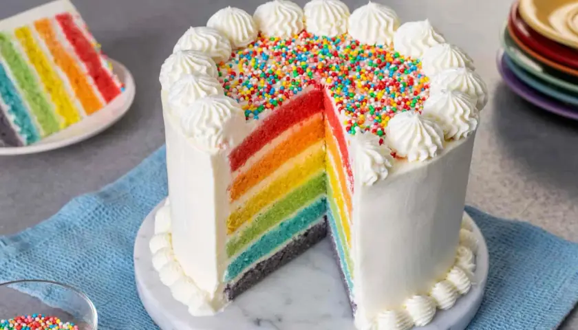 Torta arcobaleno con strati colorati, glassa bianca e zuccherini. Una fetta mostra i colori vivaci, come un vero arcobaleno.