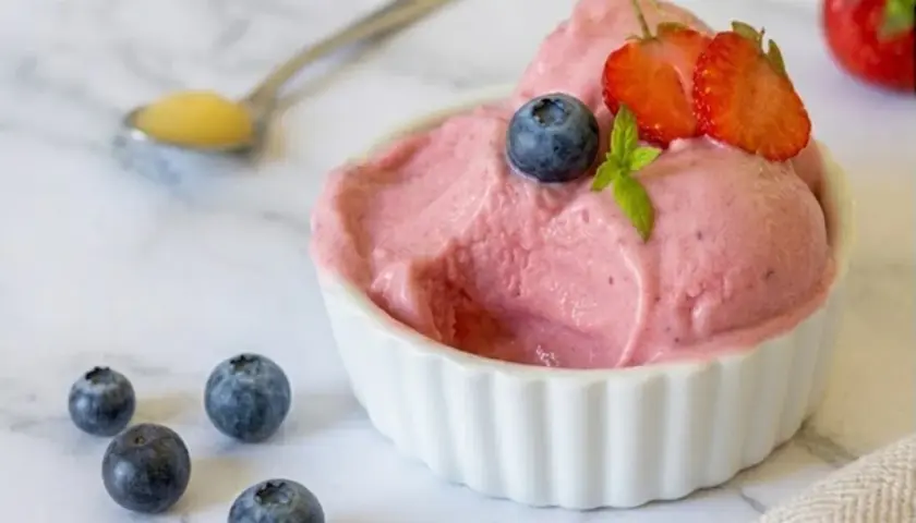 Delizioso gelato rosa, guarnito con mirtilli e fragole, servito in una ciotola bianca. Intorno, altri mirtilli e un cucchiaino con un po' di miele. Perfetto per un momento di fresco piacere, questo gelato palline di ghiaccio è un sogno.