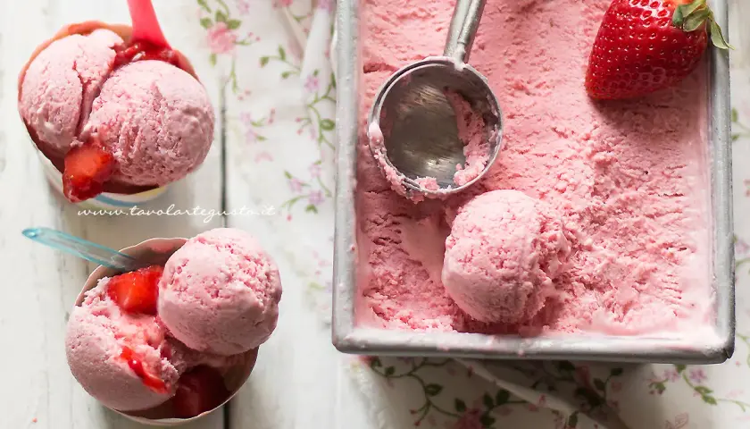 Coppette e vaschetta piene di cremoso gelato alla fragola, con fragole fresche e un cucchiaio pronto per servire.