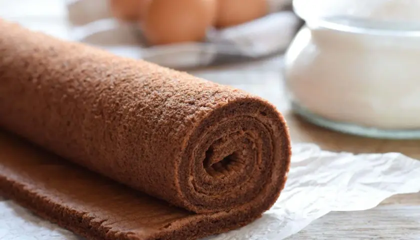 Rotolo di pasta biscotto al cacao, pronto per essere farcito. Ingredienti freschi come uova e zucchero sullo sfondo.