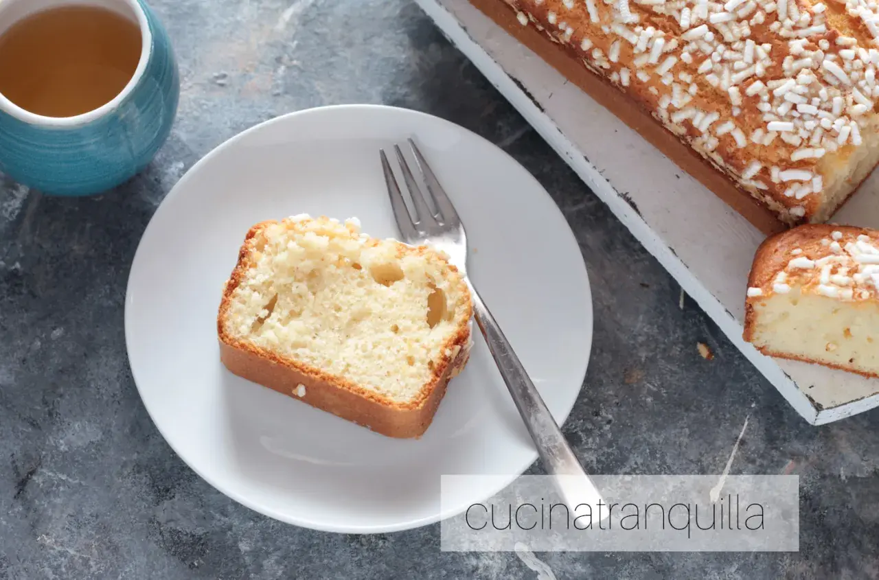 Una fetta di plumcake yogurt greco, soffice e cosparso di zuccherini, attende su un piatto bianco, accanto a una tazza di tè.