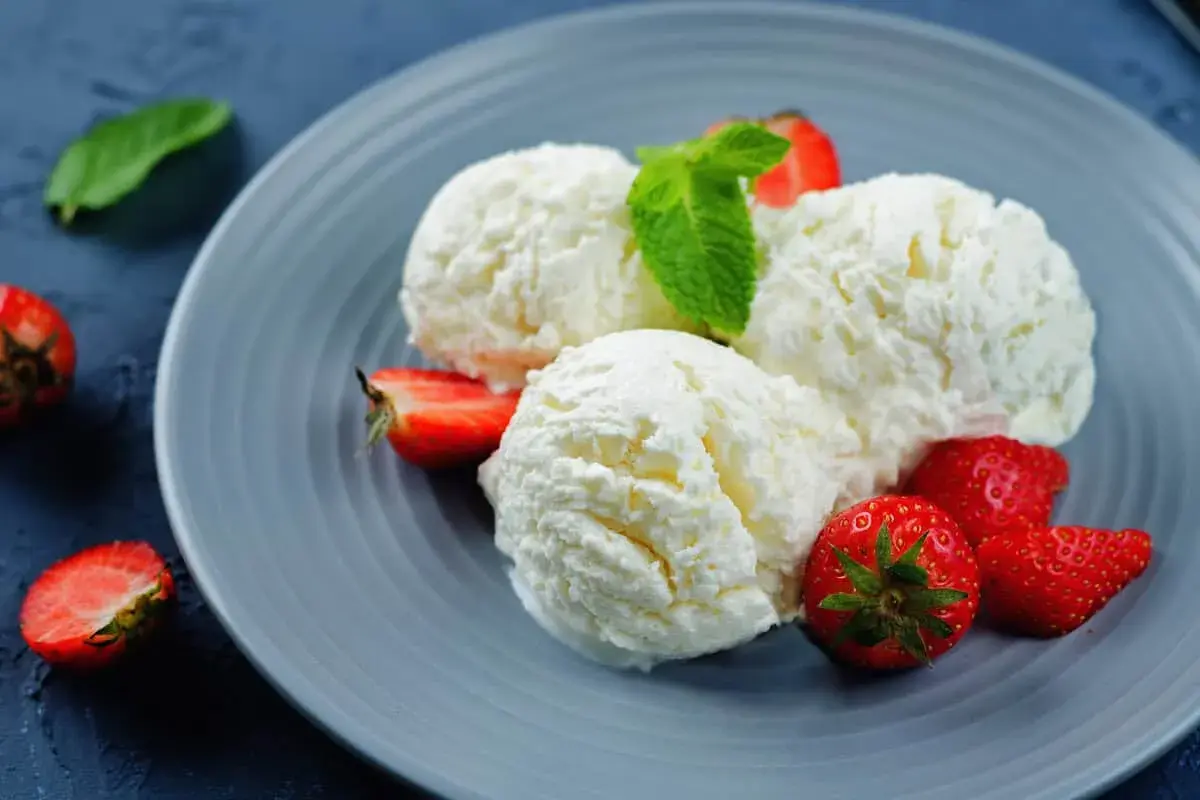 Tre palline di cremoso gelato alla ricotta, guarnite con fragole fresche e una fogliolina di menta, su un piatto grigio.