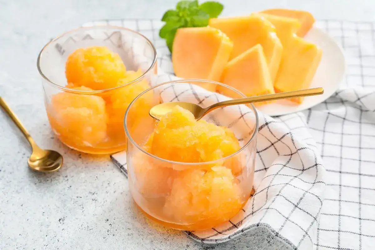 Sorbetto melone fresco e dissetante, servito in coppette di vetro con cucchiaini dorati. Fette di melone e menta sullo sfondo.