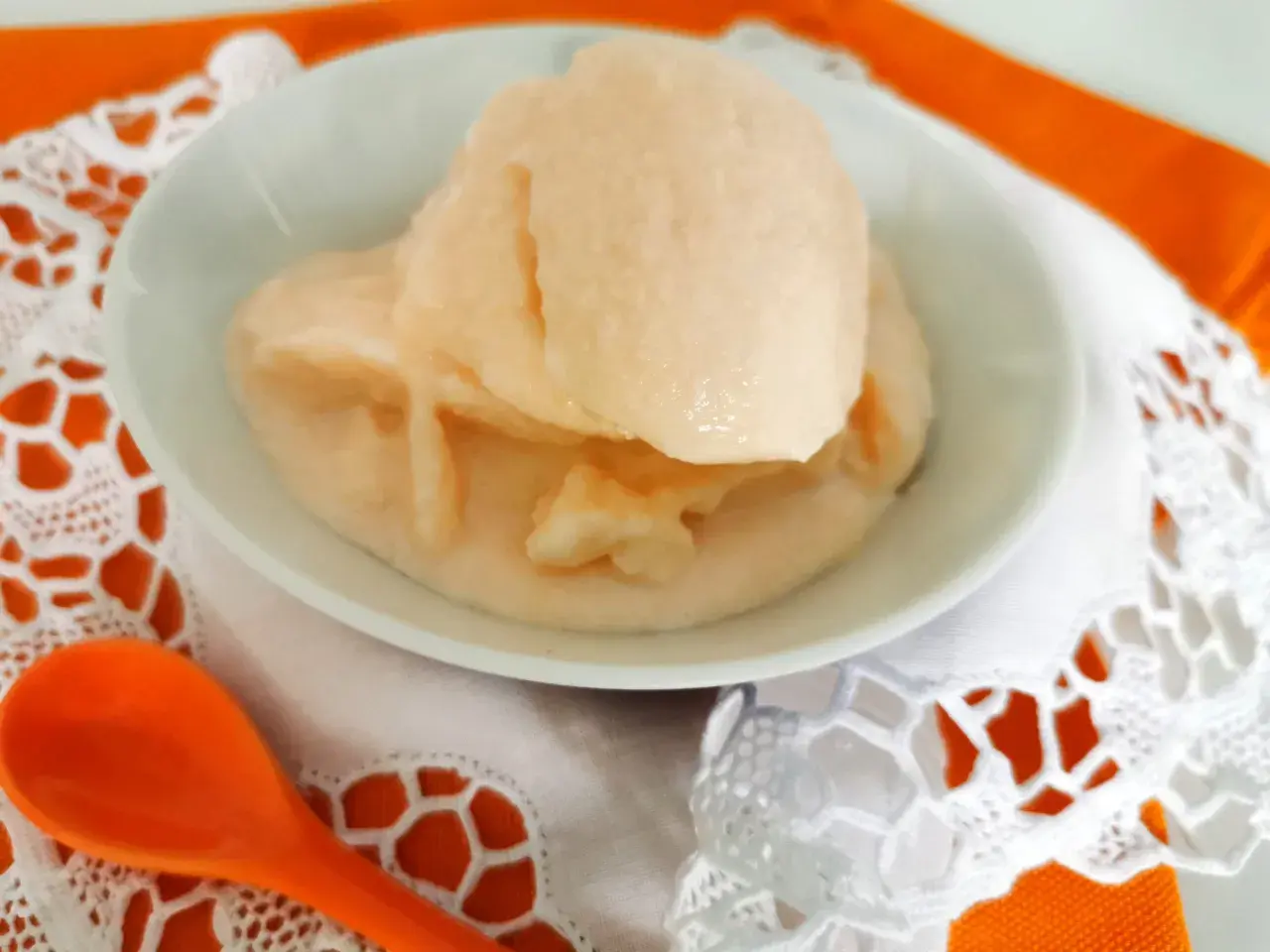 Delizioso gelato al melone solo frutta, servito in una ciotola bianca su un tovagliolo di pizzo bianco e un runner arancione. Un cucchiaio arancione è posizionato a sinistra.