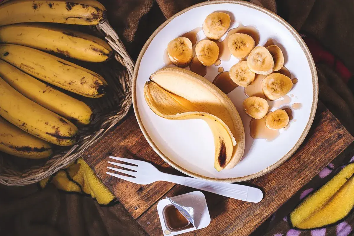 Un cestino di banane mature accanto a una banana sbucciata e affettata su un piatto, con sciroppo. Un'idea per sostituire lo zucchero con la banana.